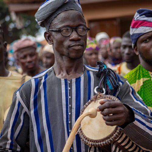 Homme en tenue rayée et bonnet traditionnel jouant d'un petit tambour, au milieu de la foule.