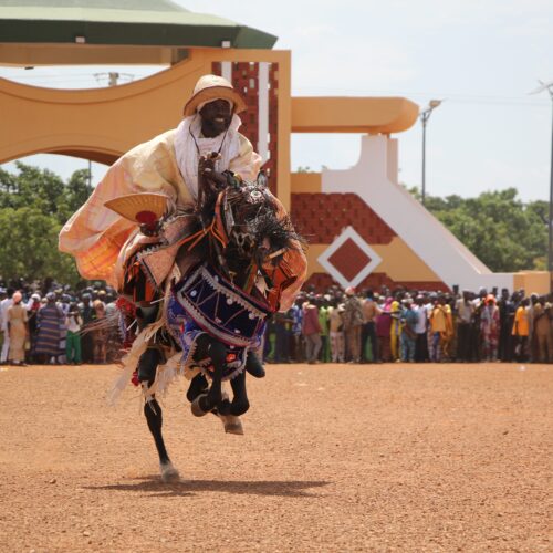 Cavalier en tenue blanche et dorée sur son cheval cabré, devant le monument de Nikki, sous les yeux de la foule.